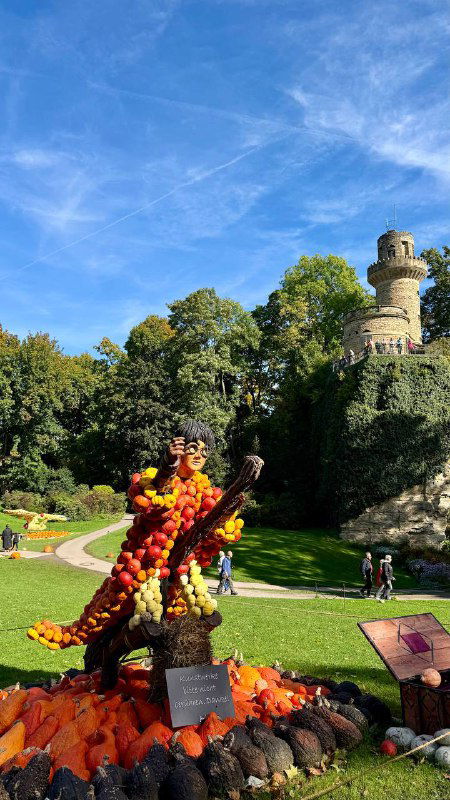 Высокая скульптура из множества оранжевых тыкв на зеленой лужайке рядом с замковой башней и деревьями — эффектная композиция фестиваля на открытом воздухе.