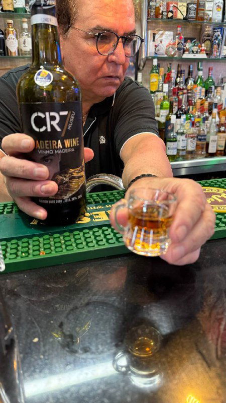 Владелец бара протягивает дегустационный шот мадеры CR7, видно бутылку с этикеткой и барную стойку с рюмкой пончи.