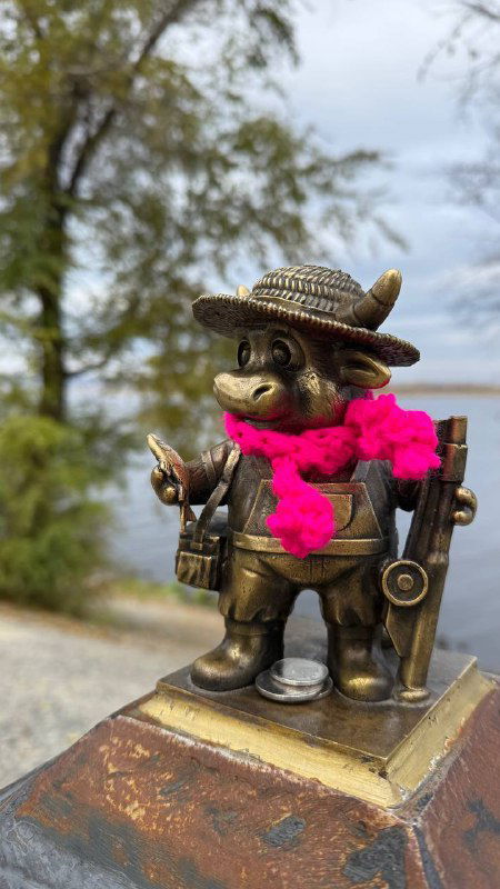 Бронзовый бычок в шляпе и с ярким розовым шарфом на грудке, установлен на каменной подставке среди осенней листвы в парковой зоне.