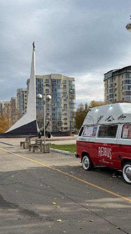 Красный фудтрак Redbus и колесо обозрения на открытой площадке набережной, городской вид с лавочками и современными зданиями на заднем плане.