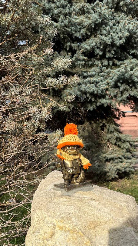 Маленькая бронзовая фигурка бычка в оранжевой шапочке, установленная на камне у зелёной хвойной ели в городском дворе Энгельса.