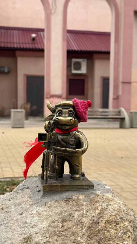 Бронзовый бычок в розовом и красном шарфе установлен у корпуса здания с колоннами — крупный план фигурки на фоне городской архитектуры.