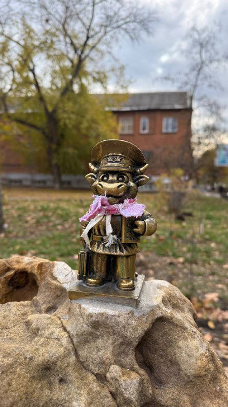 Крупный план бронзового бычка в цилиндре и с аксессуарами, стоит на камне в осеннем городском парке — видно траву и деревянные элементы.