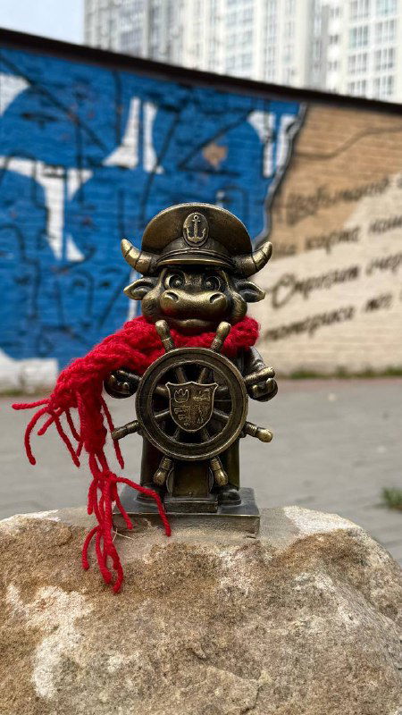 Бычок‑морячок с штурвалом и ярким красным шарфом установлен на камне на фоне граффити и городской стены в центральной части Энгельса.