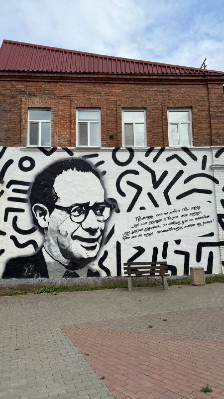 Чёрно‑белое муральное изображение портрета на стене дома в центре города, окружение с лавочкой и тротуаром — уличное граффити Энгельса.