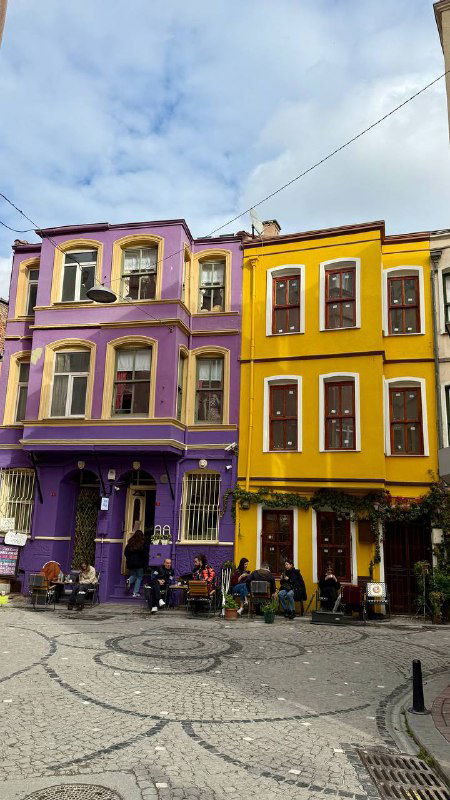Площадка с фиолетовыми и жёлтыми домами, уличными столиками и прохожими — типичная сценка радужного уголка Балата для прогулок и фото.