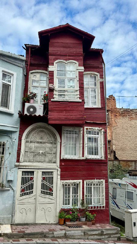 Узкий дом с деревянным фасадом тёмно-красного цвета и декоративной резьбой возле городской улицы — пример необычного жилого здания района.