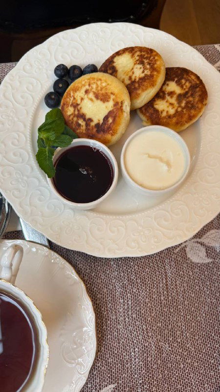 Тарелка с тремя золотистыми сырниками, рядом мисочки с ягодным соусом и сметаной, на фоне чашки чая и кружевной салфетки — аппетитная порция.