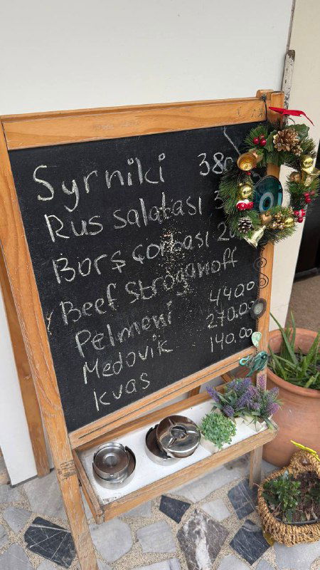 Меловая доска-меню у входа с надписями на русском языке и ценами, украшенная рождественским декором — подчёркивает домашний формат заведения.