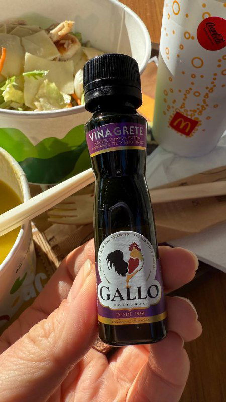 Рука держит мини‑бутылочку виноградного уксуса Gallo на фоне миски с салатом и бумажного стакана из ресторана Макдональдс в Португалии.