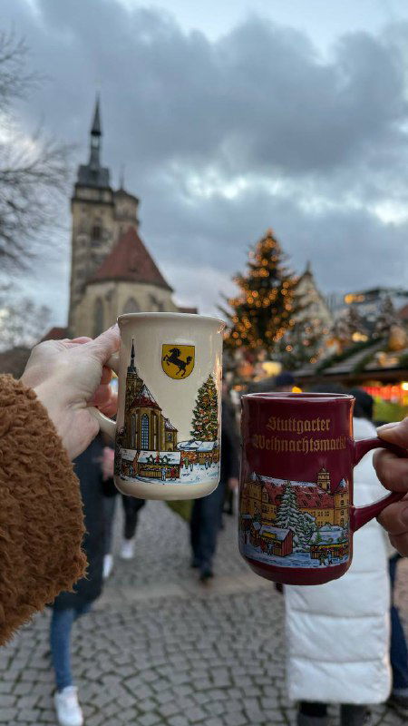 Две декоративные керамические кружки с рождественскими изображениями на фоне ёлки и декора ярмарки, видно движение людей и зимнюю атмосферу.