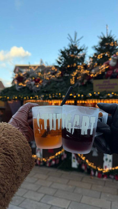 Две пластиковые кружки с разноцветными напитками и рождественскими силуэтами на переднем плане, на заднем плане ярмарочная подсветка и елочные гирлянды.