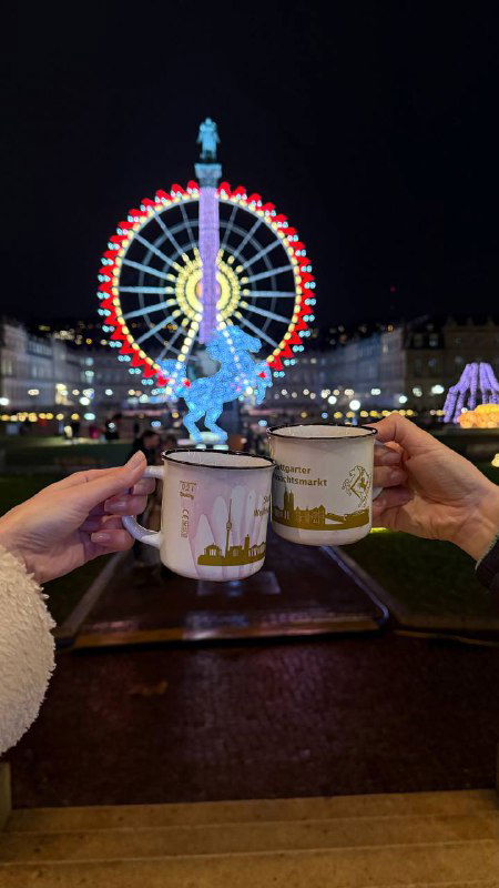 Ночная сцена: два человека держат белые кружки с тёмным напитком на фоне ярко освещенного колеса обозрения и праздничных огней на площади.