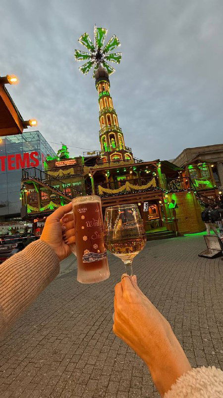 Руки подносят бокал пива и бокал белого вина на фоне ярмарочной карусели и украшений — контраст кружек и других праздничных напитков на ярмарке.