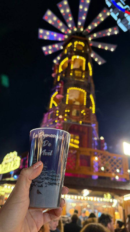 Крупный план тёмной цилиндрической кружки с надписью на переднем плане и размытой высокой ярмарочной аттракцией в огнях на заднем плане вечером.