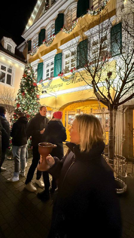 Человек держит кружку с глинтвейном у украшенного желтыми фасадом дома; вокруг толпа людей, огни и рождественские украшения создают уютную атмосферу.