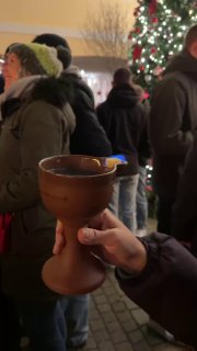 Крупный план деревянного кубка с горячим напитком в руках посетителя ярмарки; вокруг тёплые огни, люди и атмосферные декорации праздника.