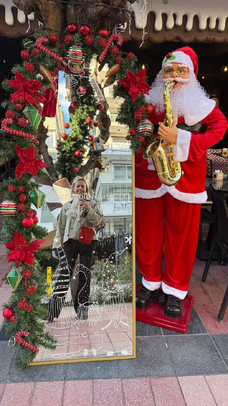 Ростовой Санта и зеркальная фотозона у кафе: человек позирует рядом с фигурой Санты и венком — уличная праздничная сцена в Измире.