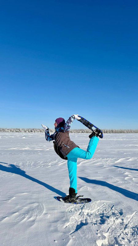 Участница в тёплой одежде на снегоступах делает растяжку на широкой заснеженной равнине под чистым голубым небом, простор вокруг.