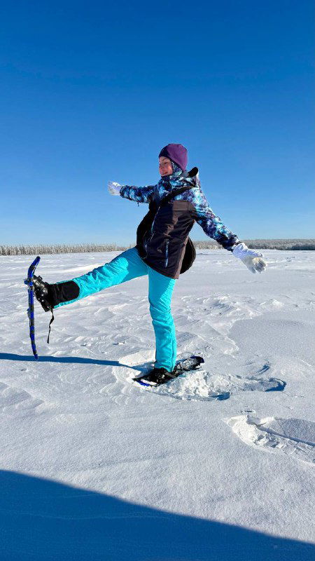 Женщина в бирюзовых штанах на снегоступах шагает по глубокому снегу, поднятая рука и яркое небо создают динамичный кадр зимней прогулки.