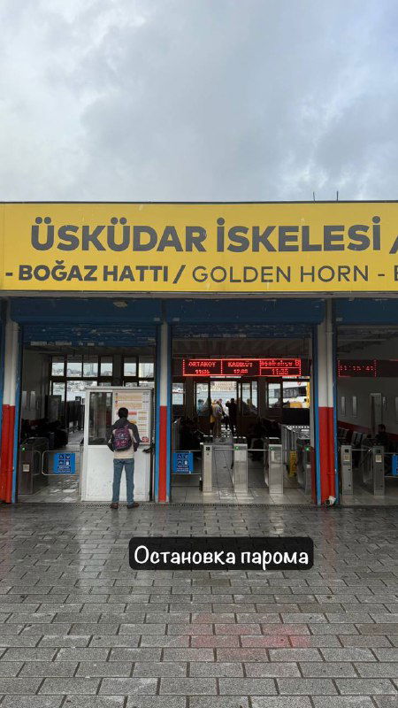 Входная зона паромной остановки Üsküdar İskelesi в Стамбуле: жёлтая табличка с указанием маршрута Bosphorus/Golden Horn и люди под дождём