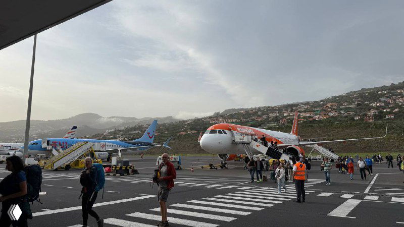 Пассажиры идут по перрону рядом с небольшими самолётами в аэропорту Мадейра; видна горная местность и взлётно-посадочная полоса у побережья.