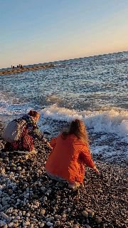 Две девушки на галечном пляже у кромки воды; волна поднимается и захватывает берег, видны прибой, дальний берег и вечернее небо