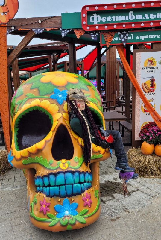 Яркая большая инсталляция черепа в стиле «День мёртвых», на ней сидит ребёнок в тёплой одежде, вокруг — фестивальные ларьки.