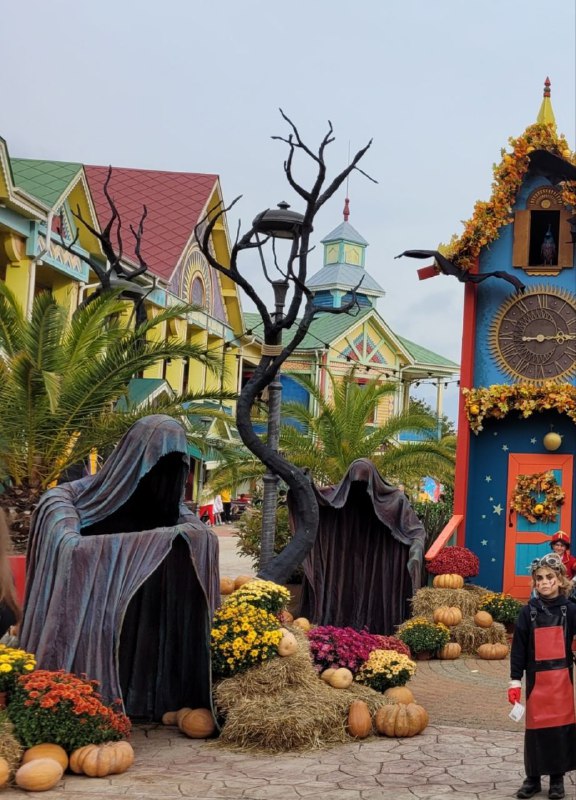 Две мрачные фигуры в чёрных плащах и голыми деревьями на фоне жанровой архитектуры парка, украшенного осенними цветами и элементами хэллоуина.