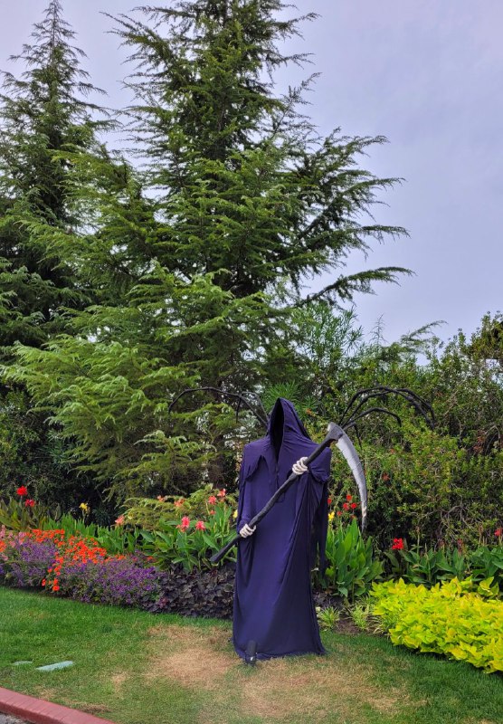 Человекообразная фигура с косой в тёмном одеянии стоит на газоне в окружении цветущих клумб и декоративных растений, вечерняя иллюминация.