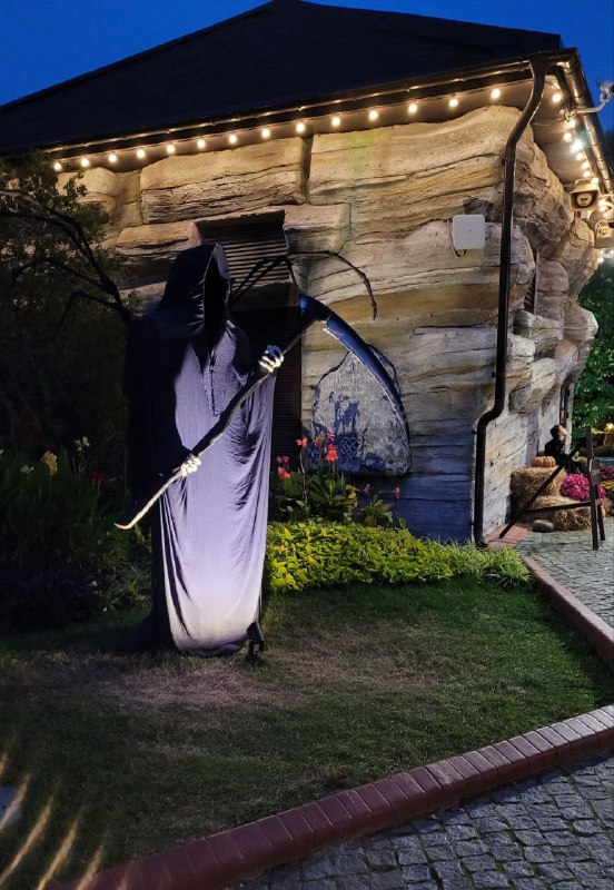 Силуэт фигуры с косой у домика в сумерках, расставленные тыквы и приглушённое освещение создают зловещую, но эстетичную сцену.