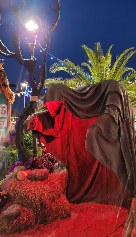 Фигура в плаще, подсвеченная красным светом, рядом декоративные тыквы и снопы соломы — ночная сцена хэллоуинского фестиваля в парке.