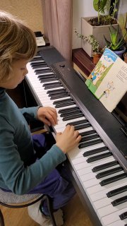 Ребёнок за цифровым пианино дома, крупный план рук на клавишах; на стойке открыт нотный сборник с цветной детской иллюстрацией.