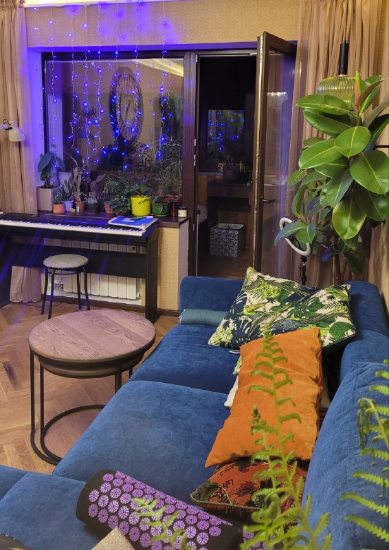 Уютный уголок с синим освещением, диван с подушками, журнальный стол и растения; гирляндные огни отражаются в стеклах окна.