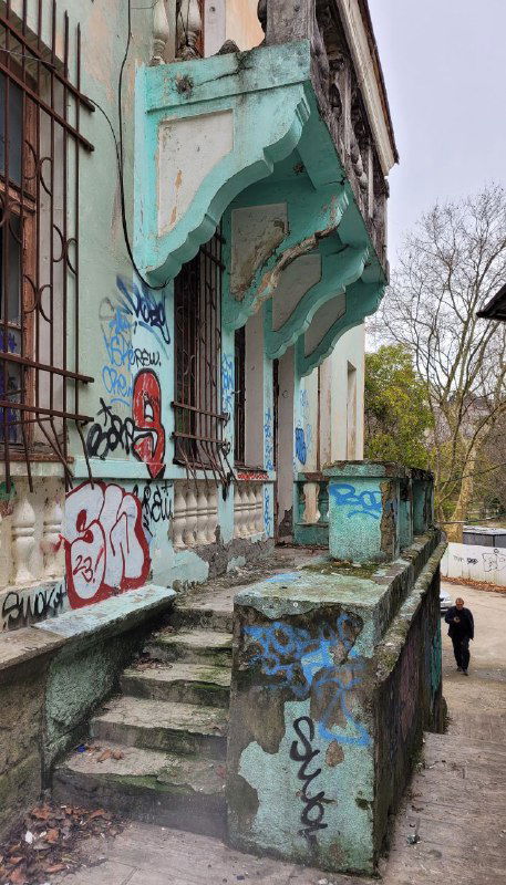 Старая многоэтажка с граффити, облупившейся лепниной и бетонной лестницей; городской пейзаж Сочи в пасмурный день.