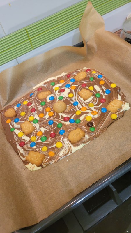 Вид сверху на большую плитку из шести разных шоколадок с печеньем и M&M's на пергаменте, запечённую в духовке и украшенную белой и темной глазурью.