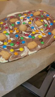 Крупный план украшенной шоколадки с печеньем и цветными драже M&M's на пергаменте, видна текстура шоколада и детали оформления на кухонной поверхности.