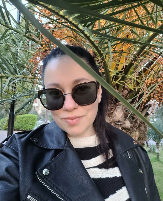 Селфи человека в тёмных очках на фоне пальмы с яркими оранжевыми плодами; видны кожаная куртка и полосатый свитер, листья вокруг.
