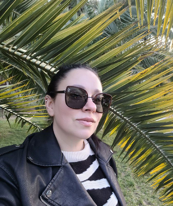 Селфи под листьями пальмы: лицо в очках и часть плеча, сзади — кисти оранжевых плодов и городской зелёный фон.