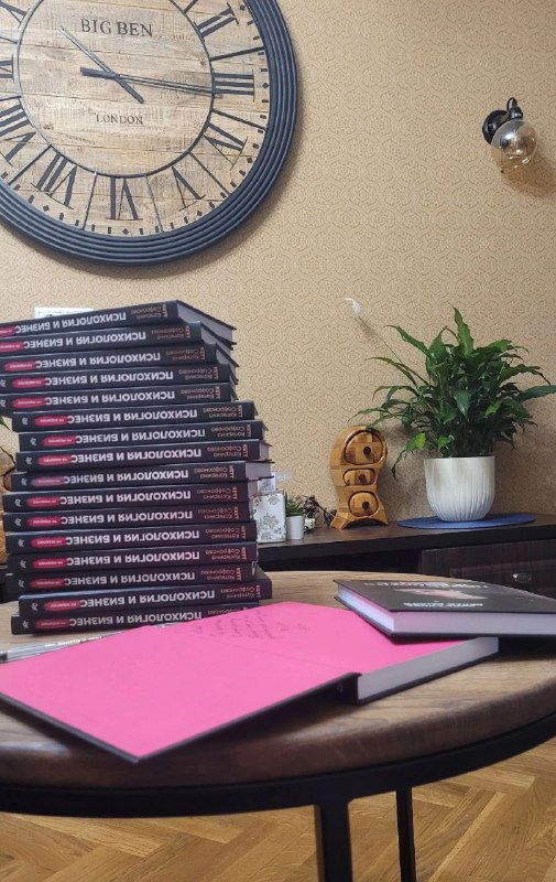 Стол с аккуратной стопкой книг, настенные часы, блокнот и растение — рабочая композиция, демонстрирующая тираж книги и рабочее пространство.