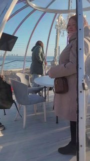 Гость в тёплом пальто внутри прозрачного купола на фоне моря и набережной; атмосфера уличного ужина в тепле купола.