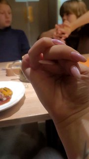 Крупный план руки с плетёной «косичкой» из пальцев за столом в гастро-баре, на заднем плане размытые люди и тарелка с едой.