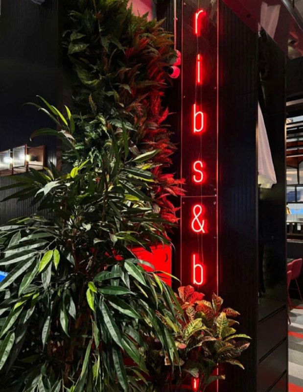 Интерьер кафе с декоративными зелёными растениями на переднем плане и вертикальным неоновым знаком у входа, тёплая красная подсветка.