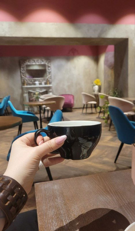 Рука с чашкой какао в первом плане на фоне столов и кресел кафе; стильный интерьер с мягкой подсветкой и спокойной палитрой.