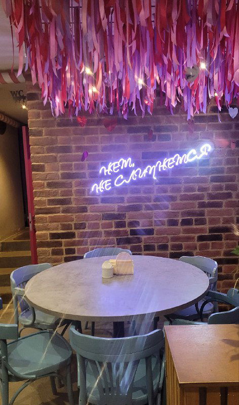 Неоновая надпись на стене кафе «Нет, не слипнется!» в розово-фиолетовой гамме, столик и стулья под декоративной драпировкой из ленточек.