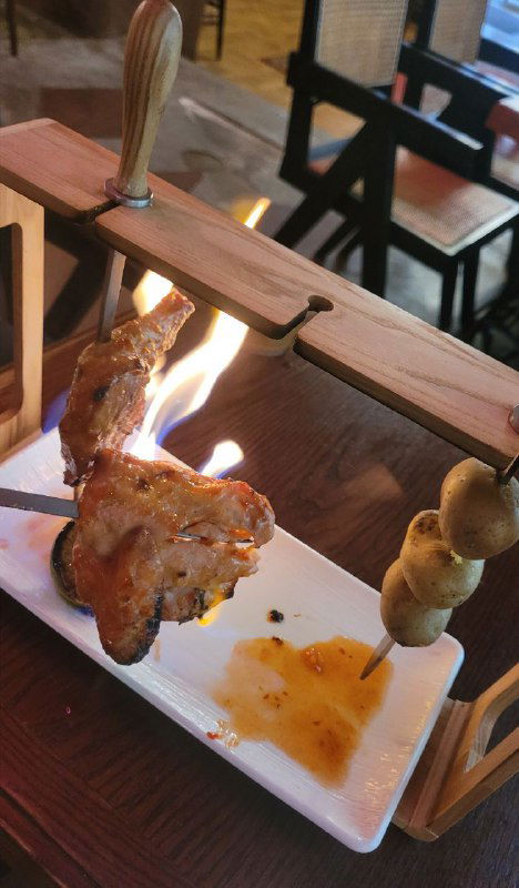 Фрагмент: кусок шашлыка на шпажке, подвешенный к деревянной перекладине над пламенем — видно капли соуса и языки огня.