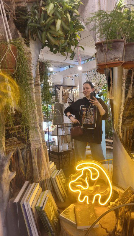 Отражение зеркала и полки с книгами и сувенирами, внутри виден владелец пространства и неоновая вывеска в форме логотипа.