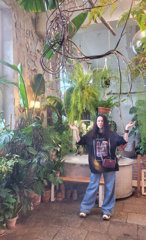 Человек в чёрной толстовке стоит среди подвешенных растений и больших вазонов — атмосферное фото на фоне зелёной стены.