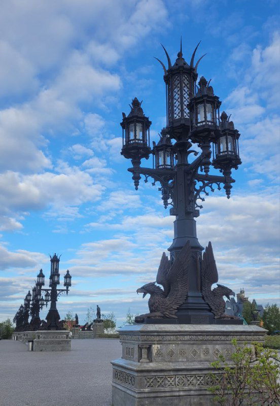 Несколько фонарных столбов с бронзовыми скульптурами на открытой набережной, облачное небо создаёт глубину кадра