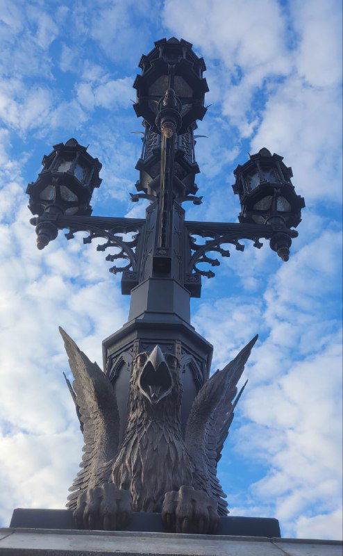 Фрагмент фонарного столба с декоративным грифоном и сложной кованой решёткой на фоне облачного неба